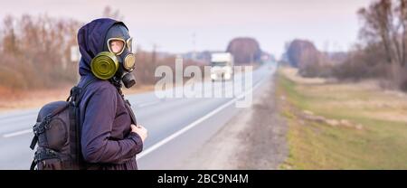 Une fille dans un masque à gaz noir se tient au bord d'une autoroute de banlieue. Bannière. La fille essaie d'arrêter de passer des voitures afin de quitter la ville à qui Banque D'Images