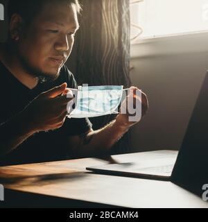 Un homme asiatique dans un masque médical travaillant sur un ordinateur portable dans la zone sombre. Masque de port. Banque D'Images
