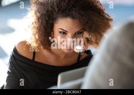 Gros plan portrait d'une jeune femme souriante en robe noire et avec des cheveux brun frisé, propulse son visage avec sa main et regardant les ordinateurs portables Banque D'Images