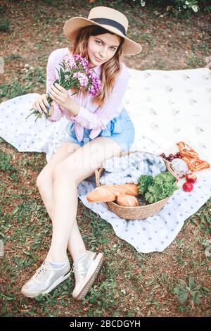 belle fille détendue dans un chapeau de paille dans la nature pique-nique panier de fleurs dans les rayons du doux coucher de soleil, camping pique-nique. Banque D'Images