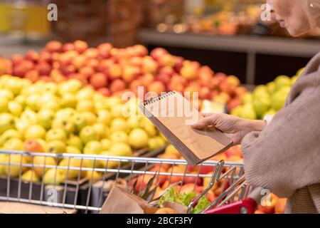 Femme mûre consommateur avec bloc-notes sur les produits alimentaires dans le panier en regardant dans la liste d'achats tout en se déplaçant le long de l'affichage des fruits Banque D'Images