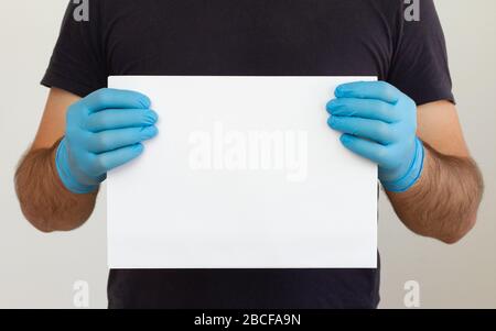 Un homme dans un t-shirt noir et des gants médicaux contient une feuille de papier blanche. Maquette. Coronavirus. Quarantaine. Avertissement Banque D'Images