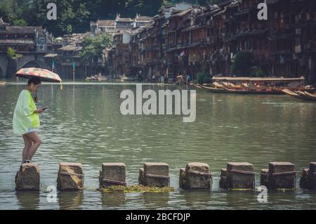 Feng Huang, Chine - Août 2019 : dame chinoise avec un parapluie et téléphone mobile traversant les eaux sur des pierres de pas sur la rivière Tuojiang, coulant des eaux Banque D'Images
