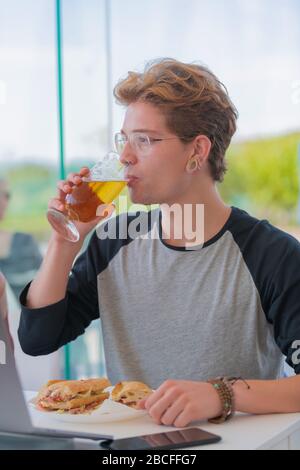 jeune homme buvant de la bière assis devant une table dans laquelle il y a quelques morceaux de sandwich avec un ordinateur portable. Image verticale. Banque D'Images