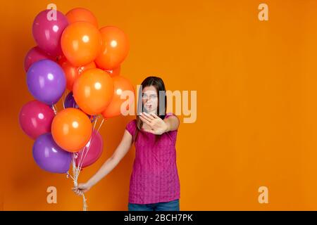 belle femme décontente dans un t-shirt rose avec des ballons d'air colorés lumineux montrant un geste d'arrêt isolé sur fond orange. fête d'anniversaire. espace pour le texte Banque D'Images