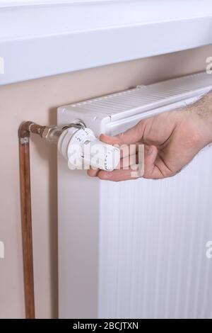 La main de l'homme maintient le distributeur de température sur le radiateur et définit la valeur requise. Radiateur blanc et tuyaux d'eau en cuivre sur la seconde ba Banque D'Images