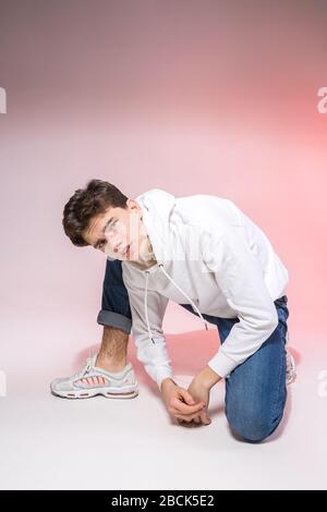 Tendance beau jeune modèle européen masculin vêtu d'un sweat-shirt blanc, d'un Jean bleu et de sneakers blanches se posant en studio sur un fond rose pour Banque D'Images