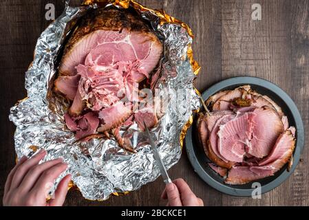 La main de la femme coupe des morceaux de jambon d'une spirale coupée de jambon glacé et cuit dans un emballage en feuille sur une table en bois, plaque noire avec des tranches Banque D'Images