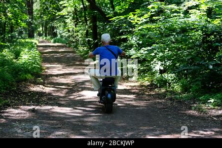 L'homme adulte conduit son scooter électrique dans la ruelle du parc un jour d'été parmi de nombreux arbres verts. Banque D'Images