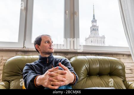 Jeune homme assis sur un canapé dans un appartement plat avec vue sur Varsovie, Pologne célèbre Palais de la Science et de la Culture bâtiment à travers la fenêtre Banque D'Images