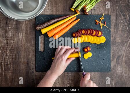 Les mains de la femme coupantes les carottes arc-en-ciel sur une planche à découper noire, un bol en acier inoxydable, une table en bois Banque D'Images