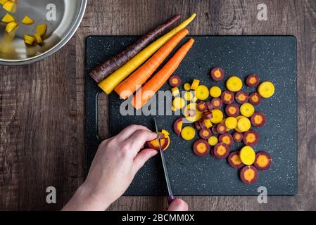 Les mains de la femme coupantes les carottes arc-en-ciel sur une planche à découper noire, un bol en acier inoxydable, une table en bois Banque D'Images