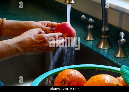 Une femme lave/désinfecte des fruits de pomme frais dans l'évier de la cuisine avec un lavage à l'eau sur la surface. Des gouttelettes s'accrochent aux agrumes dans le panier nex Banque D'Images