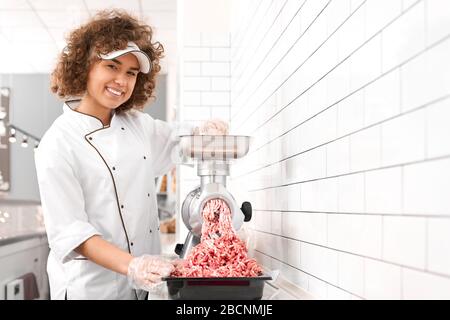 Jeune femme avec des cheveux bouclés mettant de petits morceaux dans le moulin à viande et les grids dans la viande hachée. Une femme plutôt souriante qui travaille dans la boucherie, regarde Banque D'Images