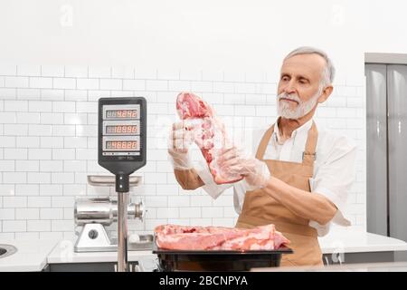 Foyer sélectif de boucher mâle senior tenant un morceau de viande fraîche brute rouge dans les mains. Homme joyeux derrière le comptoir du magasin montrant la viande, bol du réfrigérateur Banque D'Images