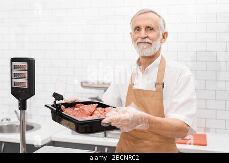 Portrait d'un boucher masculin souriant tenant un morceau de viande fraîche brute rouge dans les mains. Homme joyeux derrière le comptoir du magasin montrant la viande, mettant le bol Banque D'Images