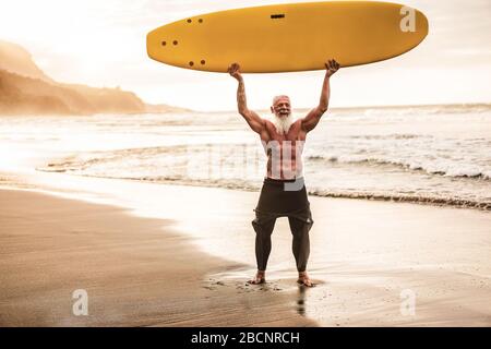 Surfeur expérimenté en tatouage qui tient planche à surf sur la plage au coucher du soleil - un vieil homme heureux qui s'amuse à faire du sport extrême - un concept de personnes âgées joyeuses - se concentrer sur la haute Banque D'Images