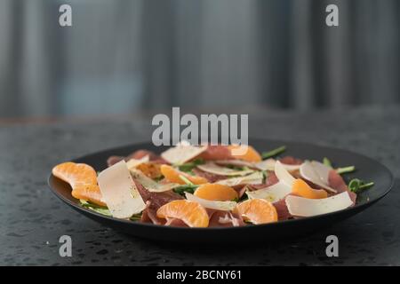 Salade avec prosciutto, mandarines et fromage d'époque sur une assiette noire Banque D'Images