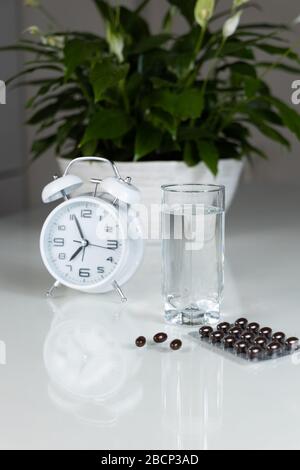 Réveil blanc et pilules médicales avec un verre d'eau sur la table. Santé et médecine. Vitamines et compléments minéraux en capsules. Banque D'Images