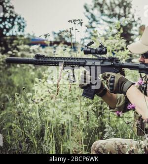 Un militaire ou un joueur aérodoux dans un costume de camouflage est assis dans l'herbe et vise d'un fusil automatique sur le côté Banque D'Images