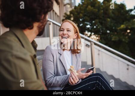 Portrait de la jeune femme gaie en conversation avec l'homme pendant la pause tout en mangeant un sandwich pour le déjeuner Banque D'Images