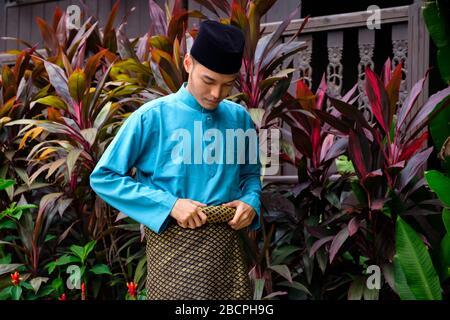 Malay homme portent un tissu traditionnel et ajuste son songket samping juste à l'extérieur de sa maison traditionnelle en bois. Banque D'Images