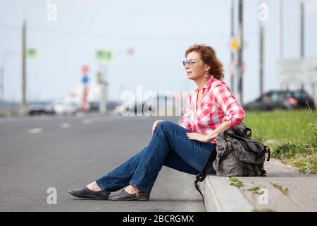 Femme caucasienne mûre dans des lunettes assis sur le trottoir de la route et la randonnée, attendant la voiture Banque D'Images