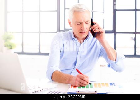 Photo d'un homme d'affaires expérimenté portant une chemise assis derrière son ordinateur portable et faisant un appel téléphonique au bureau. Banque D'Images