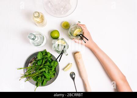 Fille tient un cocktail de mojito frais dans sa main. Ingrédients pour faire une boisson d'été sur une table blanche. Vue sur le dessus, plat Banque D'Images