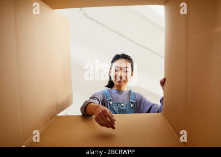 Vue en bas angle sur une femme asiatique souriante atteignant dans une boîte en carton lors de l'emballage ou du déballage pour une nouvelle maison, espace de copie Banque D'Images