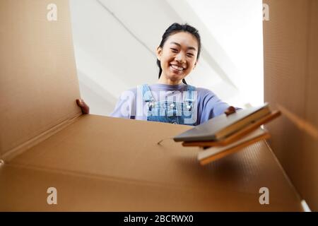 Vue à bas angle sur la jeune femme asiatique regardant dans une boîte en carton et souriant heureux tout en emballant ou en déballant pour la nouvelle maison, espace de copie Banque D'Images