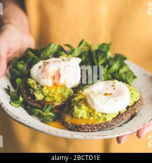 Oeuf poché et avocat sur pain de seigle toast sur plaque dans les mains femelles. Petit déjeuner ou repas sains. Cuisine gastronomique Banque D'Images