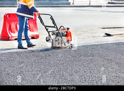 Un travailleur routier compacte l'asphalte dans une section clôturée de la route sur la chaussée à l'aide d'un compacteur vibrant à essence. Banque D'Images