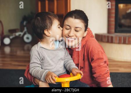 Charmant petit garçon embrassant sa mère avec des cornichons tout en jouant avec une voiture qui reste à la maison en quarantaine Banque D'Images