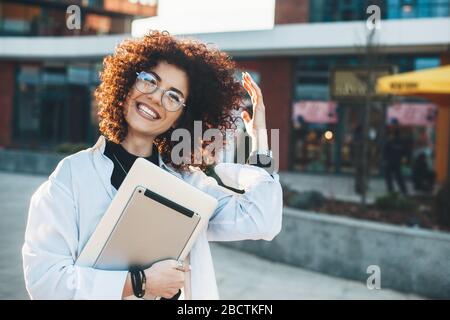 Un homme d'affaires aux cheveux bouclés se posant à l'extérieur dans des lunettes avec des gadgets modernes souriant à l'appareil photo Banque D'Images