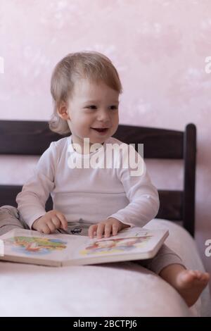 Un petit garçon aux cheveux blonds est assis sur le lit et souriant, tenant un livre dans ses mains. Un petit garçon est assis sur un lit avec un livre dans ses mains pendant Banque D'Images