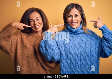 Âge moyen beau couple de sœurs portant un pull décontracté sur un arrière-plan jaune isolé souriant montrant et pointant avec les doigts dents Banque D'Images