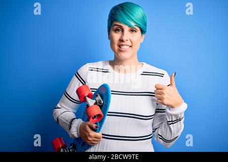 Jeune femme patineuse avec cheveux de mode bleu tenant le skateboard sur fond bleu isolé heureux avec grand sourire faisant bon signe, pouce vers le haut avec les doigts, Banque D'Images