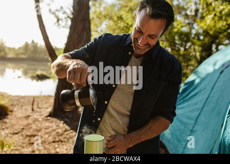 Homme souriant et mûr qui verse du café de la bouilloire dans une tasse. Homme adulte moyen ayant du café frais au camping près du lac. Banque D'Images