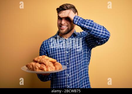 Jeune homme blond avec barbe et yeux bleus tenant plaque avec croissants français au petit déjeuner stressé avec la main sur la tête, choqué avec honte et surprise Banque D'Images