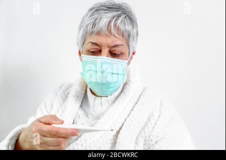 Coronavirus. Malade senior femme tenant thermomètre assis sur le lit, bouleversé vieille femme mûre attrapé froid a eu des symptômes de grippe de coronavirus à la maison seul . Banque D'Images