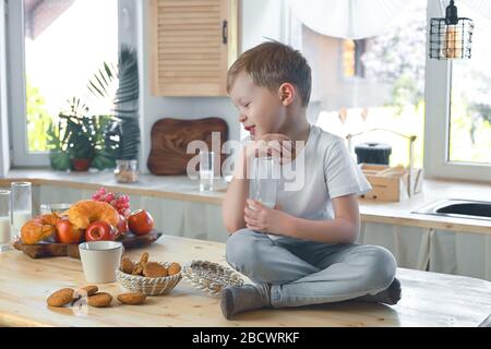 Petit garçon caucasien souriant assis sur la table de cuisine et manger des biscuits de flocons d'avoine avec du lait. Banque D'Images