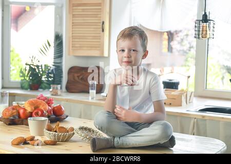 Petit garçon caucasien souriant assis sur la table de cuisine et manger des biscuits de flocons d'avoine avec du lait. Banque D'Images