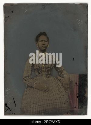 Tintype d'une jeune femme assise. Une photographie teintée à la main d'une jeune femme assise. Ce portrait représente une jeune femme assise avec son bras gauche drapé sur un accoudoir sur son côté. Portant une robe plaid, sa main droite reposant sur ses genoux, elle gaze directement à la visionneuse eh. Tintype d'une jeune femme assise Banque D'Images