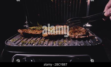 Délicieux bifteck de viande juteuse sur le gril. Viande de bœuf en marbre rôtie de qualité vieillie. Plat à rôtir électrique, poivre noir au romarin, sel. Pinces de cuisine. Banque D'Images