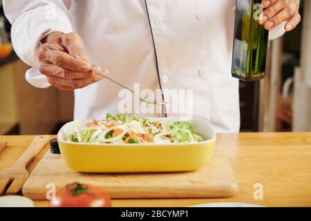 Chef verser une cuillère d'huile végétale dans un bol de salade fraîche qu'il a faite pour le déjeuner Banque D'Images
