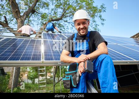 Portrait d'un technicien souriant avec tournevis électrique devant un système photovoltaïque à panneaux solaires extérieur haut inachevé avec équipe de travailleurs sur plate-forme haute. Banque D'Images