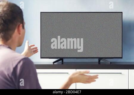 Frustré homme assis sur un canapé devant la télévision sans aucun signal. Jeune homme émotionnellement regardant la télévision et agitant ses mains dans le mécontentement. Banque D'Images