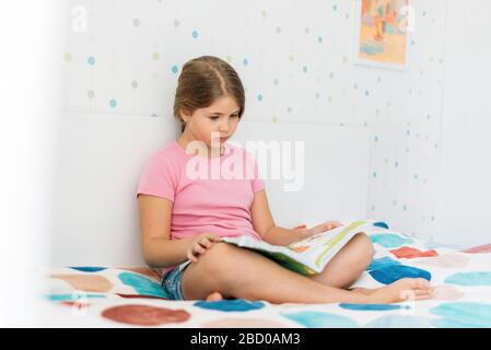 Jolie fille Tween dans un livre de lecture de t-shirt rose assis sur le lit dans une chambre lumineuse à la maison Banque D'Images