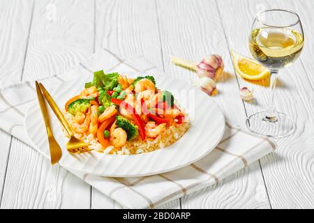 crevettes frites au poivron, à l'oignon, au brocoli et aux pois verts sur du riz cuit à la vapeur sur une plaque blanche sur une table en bois avec du vin blanc dans un verre, paysagé Banque D'Images